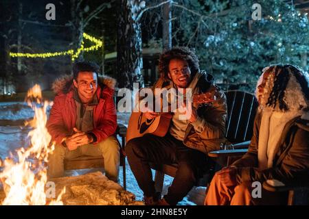 Afrikanischer junger Mann, der mit seinen Freunden Gitarre spielt, singt Lieder, die in der Nähe des Lagerfeuers sitzen, während sie sich im Winter im Wald ausruhen Stockfoto
