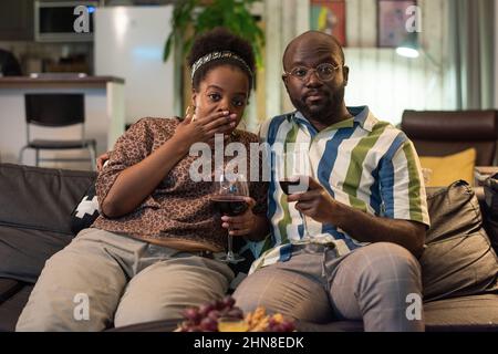 Porträt eines jungen afrikanischen Paares, das auf dem Sofa sitzt, Rotwein trinkt und zu Hause den Film genießt Stockfoto