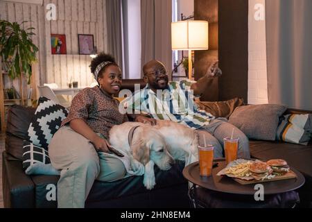 Afrikanisches Paar mit Hund, der auf dem Sofa sitzt und beim gemeinsamen Film Fast Food isst, Mann zeigt auf das fernsehen und lacht mit seiner Freundin Stockfoto