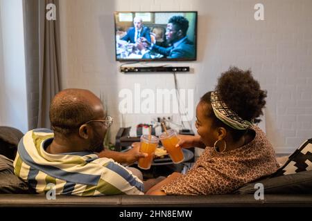 Rückansicht eines afrikanischen Paares, das mit Gläsern Saft zutoaste und lächelte, während es zu Hause auf dem Sofa saß und den Film ansah Stockfoto