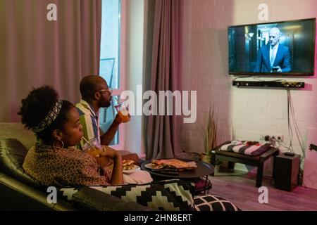 Afrikanisches junges Paar, das gemeinsam auf dem Sofa Fastfood isst und im Wohnzimmer einen Film im Fernsehen an der Wand sieht Stockfoto