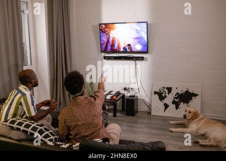 Rückansicht des afrikanischen Paares, das auf dem Sofa sitzt und mit einem breitbild-fernseher an der Wand im Zimmer Videospiele spielt Stockfoto