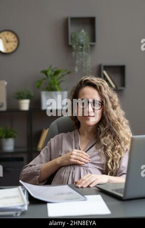 Porträt einer jungen Schwangeren in einer Brille, die die Kamera anschaut, während sie am Arbeitsplatz mit einem Laptop im Büro sitzt Stockfoto