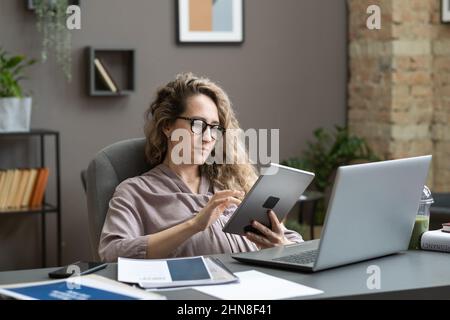 Junge, Schwangere, die online auf einem digitalen Tablet arbeiten, während sie mit einem Laptop am Tisch im Büro sitzen Stockfoto