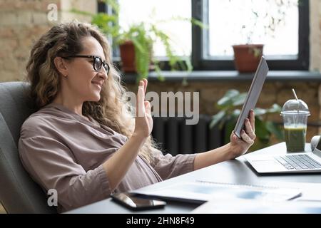 Während der Online-Konferenz im Büro winkt eine schwanger Geschäftsfrau in einer Brille zur Kamera des digitalen Tablets Stockfoto