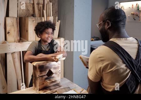 Porträt eines lächelnden afroamerikanischen Jungen, der Vater ansieht, während er in der Werkstatt die Bindung genießt Stockfoto
