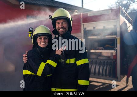 Glückliche Feuerwehrleute Mann und Frau nach der Aktion Blick auf Kamera mit Feuerwehrauto im Hintergrund Stockfoto