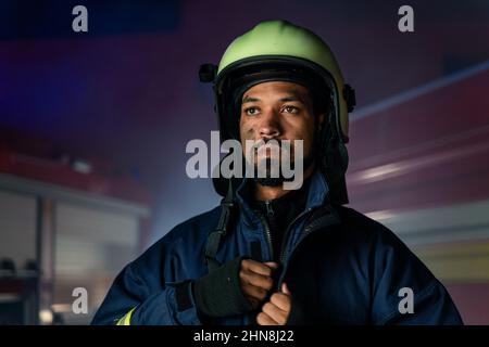 Porträt eines schmutzigen Feuerwehrmanns im Dienst mit Feuerwehrauto im Hintergrund in der Nacht. Stockfoto