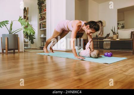 Gesunde neue Mutter, die mit ihrem Baby zu Hause arbeitet. Yogi Mama macht Push-up-Übungen auf einer Übungsmatte. Junge Mutter bonding mit ihrem Baby während sie POS Stockfoto