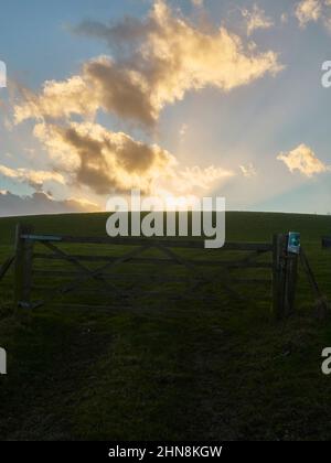 Wintersonnenstrahlen schneiden durch Wolken und über den blauen Himmel, wenn die Sonne hinter einem tief schattigen Hügel mit Tor und Grasfeld untergeht. Stockfoto