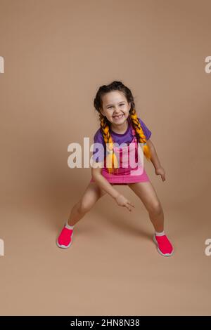 Aktive glückselige kleine Mädchen mit gelben Kanekalon Zöpfe tanzen mit Beinen weit auseinander lächelnd zeigt Stück Zeichen mit Hand in rosa Jumpsuit, lila Stockfoto