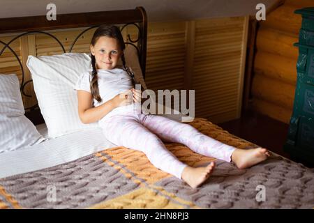 Das junge Mädchen liegt auf einem großen Doppelbett, das mit einer graugelben Decke bedeckt ist, mit einem Körper auf einem weißen Kissen und blickt auf die Kamera, die tagsüber im Holzhaus lächelt Stockfoto