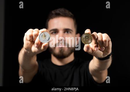 Junger bärtiger Mann mit virtueller Währung ethereum-Münze in einer Hand und Bitcoin-Münze in einer anderen auf schwarzem Hintergrund. Kryptowährung. Stockfoto