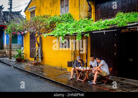 Drei Männer sitzen im Regen in der Altstadt unter einem Überhang mit grünen Pflanzen. Stockfoto