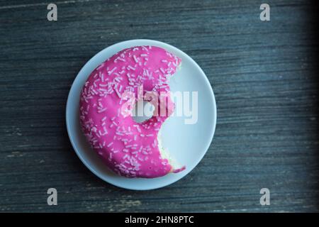 Draufsicht auf leuchtend rosa Donut auf weißem runden Teller, der zweimal abgebissen wurde. Alter Holzhintergrund. Isoliertes Objekt. Traditionelle amerikanische Süßigkeiten für Stockfoto