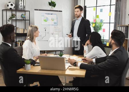 Erfolgreicher Mann in schwarzem Anzug, der in der Nähe des Flipchart steht und während der Konferenz mit multikulturellen Kollegen spricht. Eine Gruppe von Geschäftsleuten, die sich im Bürozimmer zum Briefing treffen. Stockfoto