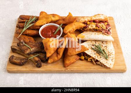 Set von Snacks: Nachos, Hähnchenflügel, gebratene Rippchen, Bohnentortillas auf einem Schneidebrett auf einem grauen Betongrund. Seitenansicht, Nahaufnahme. Stockfoto
