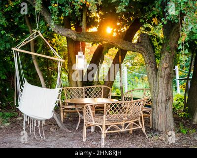 Hölzerne Gartenterrasse mit Korbmöbeln, Stühlen, Tisch und Hängematte, grüne Bäume in einem Sommersetting bei Sonnenuntergang. Sitzbereich im Garten. Kuscheliger Hygge pl Stockfoto