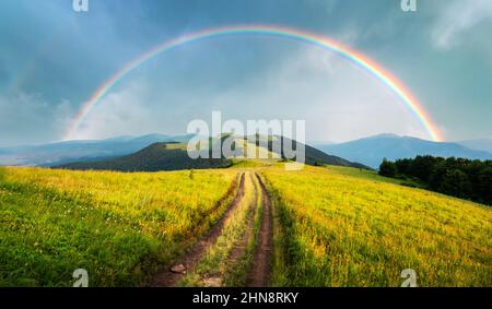 Fantastische Szene in den Bergen im Sommer. Üppig grüne Wiesen in traumhafter Abendsonne. Ländliche Straße und schöner Regenbogen in dramatischem Himmel. Landschaftsfotografie Panorama Stockfoto