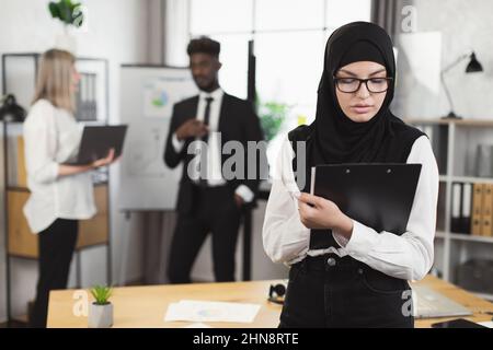 Selbstbewusste Frau in Hijab und Brillen, die auf die Zwischenablage schreibt, während ihre multiethnischen Kollegen im Hintergrund sprechen und einen Laptop benutzen. Konferenz der Mitarbeiter des Unternehmens. Stockfoto