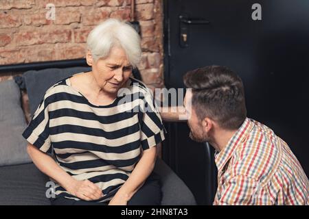 Deprimierte alte kaukasische Großmutter, die an Demenz leidet, sitzt auf einer schwarzen Couch und spricht mit ihrem erwachsenen Enkel. Hochwertige Fotos Stockfoto