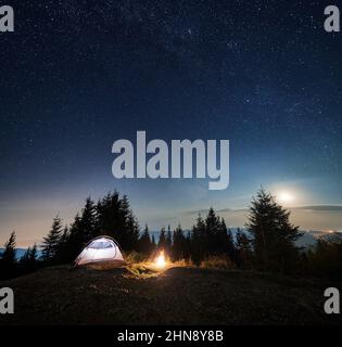 Beleuchtetes Touristenzelt auf der grasbewachsenen Spitze eines felsigen Berghügels. Kleiner Abendcampingplatz mit Zelt und Lagerfeuer vor dem Hintergrund des Waldes, Silhouetten von Bergen unter dem bunten Sternenhimmel. Stockfoto