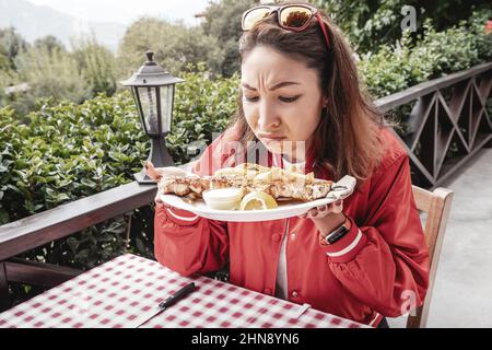 Eine Frau schnüffelt Fastfood-Gericht im Restaurant mit Ekel auf ihrem Gesicht. Das Konzept der verdorbenen Lebensmittel und Vergiftungen im Catering-Restaurant Stockfoto