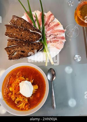 Draufsicht auf einen Teller mit Borscht mit Sauerrahm, Roggenbrotscheiben, Schweinefett und grünen Zwiebelfedern, die auf dem Tisch stehen. Stockfoto