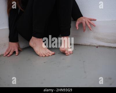 Eine unerkennbare barfuß verängstigte Frau, die in einer Ecke an der Wand sitzt Stockfoto