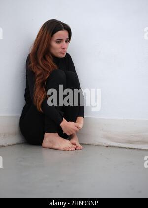 Low-Angle-Ansicht einer traurigen jungen Frau in der Ecke eines Raumes in schwarz barfuß gekleidet und am Rande der Tränen. Stockfoto