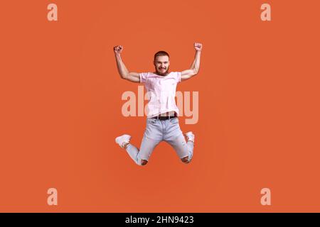 Porträt eines glücklichen, attraktiven bärtigen Mannes, der in der Luft hochgezogene Arme sprang, die Kamera ansah, Platz für Werbung kopieren und pinkes T-Shirt und Jeans trug. Innenaufnahme des Studios isoliert auf orangefarbenem Hintergrund. Stockfoto