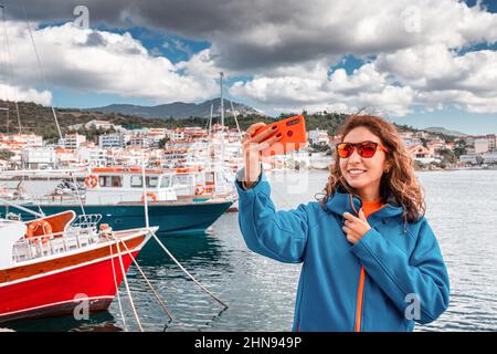 Eine Frau in einer Jacke macht ein Selfie-Foto auf einem Smartphone vor dem Hintergrund eines Piers mit Fischerbooten Stockfoto