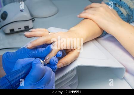 Nahaufnahme der Hände eines Maniküre-Meisters in blauen Handschuhen mit einer elektrischen Nagelfeile zum Trimmen und Entfernen von Nagelhaut in einem Schönheitssalon. Hardware-Maniküre Stockfoto