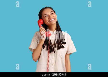 Porträt einer glücklichen lächelnden Frau mit schwarzen Dreadlocks, die ein rotes Retro-Telefon in den Händen hält, mit verträumtem Gesichtsausdruck wegschaut und ein weißes Hemd trägt. Innenaufnahme des Studios isoliert auf blauem Hintergrund. Stockfoto