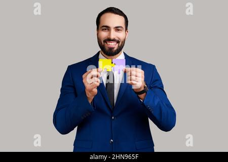 Porträt eines optimistischen bärtigen Mannes, der gelbe und violette Puzzleteile hält, Aufgaben löst, die Kamera anschaut und einen offiziellen Anzug trägt. Innenaufnahme des Studios isoliert auf grauem Hintergrund. Stockfoto
