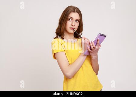 Schöne nachdenkliche Teenager-Mädchen in gelben Casual-Stil T-Shirt hält Notebook in der Hand, schriftlich Pläne für die Zukunft, denkt darüber nach, Liste zu tun. Innenaufnahme des Studios isoliert auf grauem Hintergrund. Stockfoto