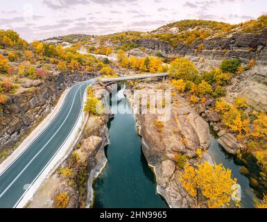Luftdrohnenansicht einer Straße, die eine enge Schlucht eines Flusses durch eine Brücke während des goldenen Herbstes in Griechenland passiert Stockfoto