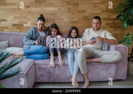 Familie mit zwei kleinen Töchtern, jeder nutzt Handy im Wohnzimmer. Stockfoto