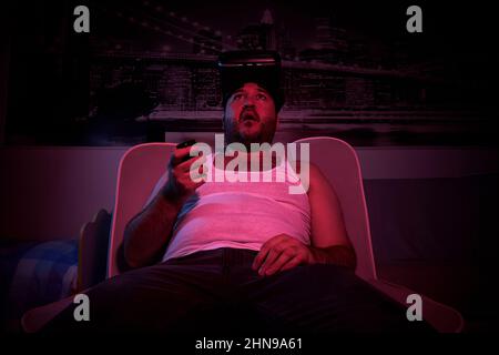 Ein 40-jähriger unrasiert übergewichtiger Mann schaut in VR-Brillen TV, zeigt verschiedene Emotionen, ist überrascht. Fauler Mann mittleren Alters Stockfoto