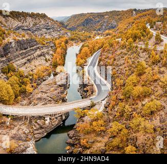 Luftdrohnenansicht einer Straße, die eine enge Schlucht eines Flusses durch eine Brücke während des goldenen Herbstes in Griechenland passiert Stockfoto