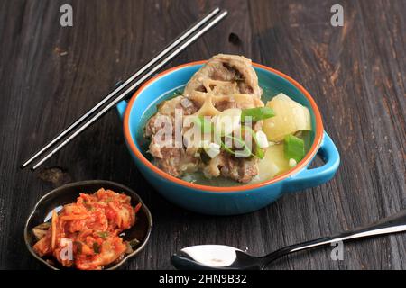 Gori Gomtang (Sokkori Gomtang) oder koreanische Rinderoxtail-Eintaufsuppe, serviert in der Blauen Schüssel mit Kimchi und geschnittenen grünen Zwiebeln, auf dem schwarzen Holztisch Stockfoto