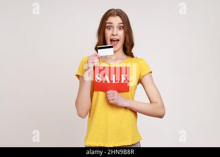 Fröhlich aufgeregt Frau in gelben T-Shirt halten zeigt Kreditkarte und Karte mit Verkauf Inschrift, bezahlen für Einkäufe mit Kreditkarte, Banking. Innenaufnahme des Studios isoliert auf grauem Hintergrund. Stockfoto