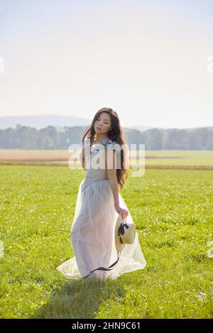 Junge schöne lange schwarze Haare asiatische Frau trägt ein lila romantisches Kleid stehen in einem hinterleuchteten Grasfeld Stockfoto