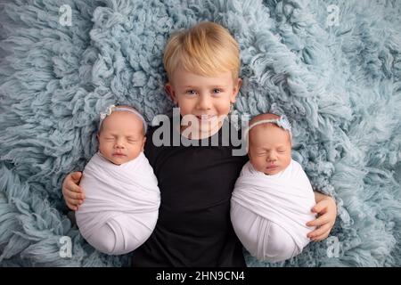 Kleine neugeborene Zwillingstündinnen in weißen Kokons auf blauem Hintergrund. Der ältere Bruder liegt zwischen ihnen und umarmt sanft seine Zwillingsschwestern. Stockfoto