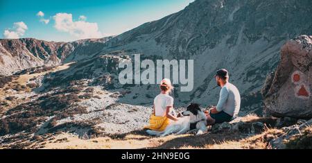 Ein Paar mit Hund sitzt auf Felsen und bewundert die Berglandschaft Stockfoto