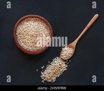 Brauner Reis in einer Holzschüssel mit Holzlöffel auf schwarzem Hintergrund, Draufsicht Stockfoto