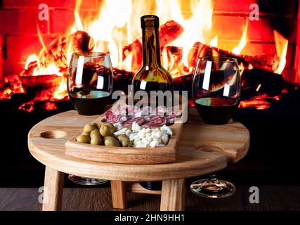 Hölzerner Weintisch mit Vorspeisen für Wein gegen elektrischen Kamin. Weingläser mit Blauschimmelkäse, Fuetwurstsalami und Oliven auf Holzbrett. Stockfoto