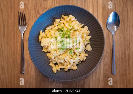 Käsespätzle in schwarzem Teller serviert mit Zwiebel und Schnittlauch mit Besteck auf Holztisch Stockfoto