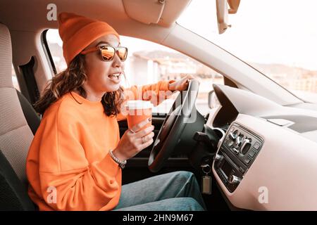 Ein Teenager-Student fährt mit einem Auto durch die Straßen der Stadt. Eine Hand ist am Lenkrad, die andere hält eine Tasse Tee oder Kaffee. Stockfoto