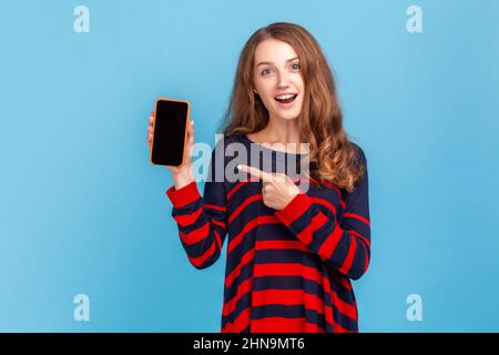 Erstaunt Frau trägt gestreiften lässigen Stil Pullover, zeigt auf Handy mit leeren schwarzen Bildschirm für Werbung, überrascht Ausdruck. Innenaufnahme des Studios isoliert auf blauem Hintergrund. Stockfoto